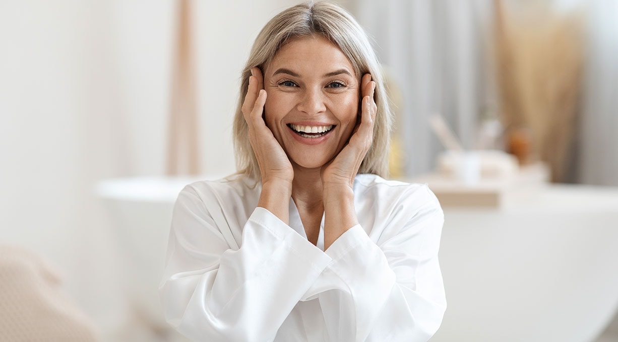 Lächelnde Frau berührt mit beiden Händen ihr Gesicht in heller Umgebung Fokus auf Hautpflege und Wohlbefinden