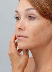 Frau mit zusammengebundenen Haaren betrachtet ihr Gesicht im Spiegel und berührt sanft ihre Wange.