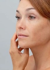 Une femme aux cheveux attachés regarde son visage dans le miroir et effleure délicatement sa joue.
