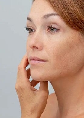 Une femme aux cheveux attachés regarde son visage dans le miroir et effleure délicatement sa joue.