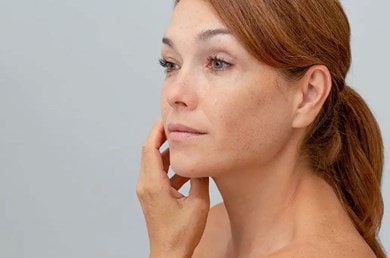 Une femme aux cheveux attachés regarde son visage dans le miroir et effleure délicatement sa joue.