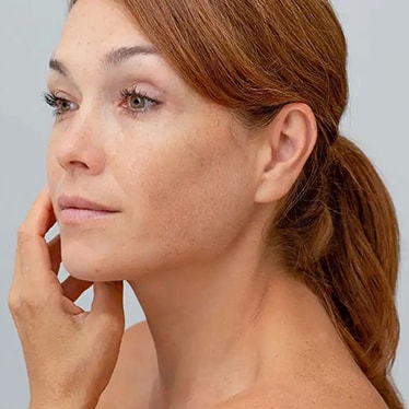 Une femme aux cheveux attachés regarde son visage dans le miroir et effleure délicatement sa joue.