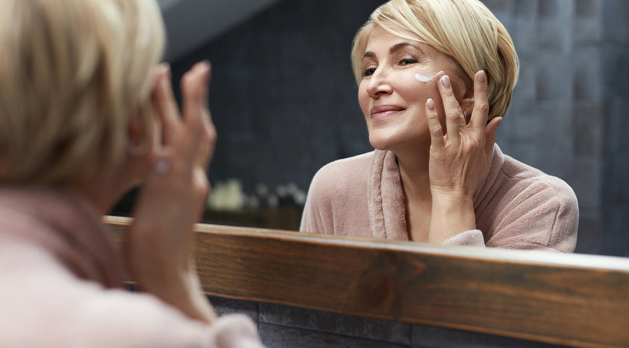 Une femme applique de la crème sur son visage devant le miroir.