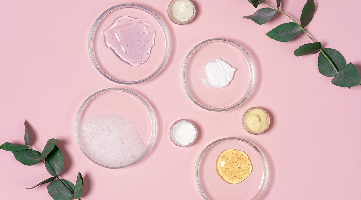 Divers produits de soin pour la peau dans des coupelles en verre sur un fond rose.