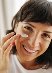 Une femme souriante applique une crème sur son visage.