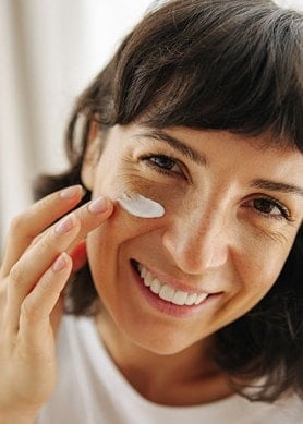 Une femme souriante applique une crème sur son visage.