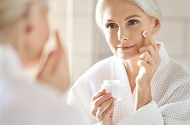 Eine Frau im Bademantel betrachtet sich lächelnd im Spiegel, während sie Creme aufträgt.