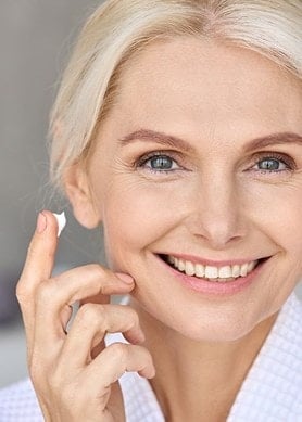 Eine lächelnde Frau hält einen Finger mit Creme nahe an ihre Wange.