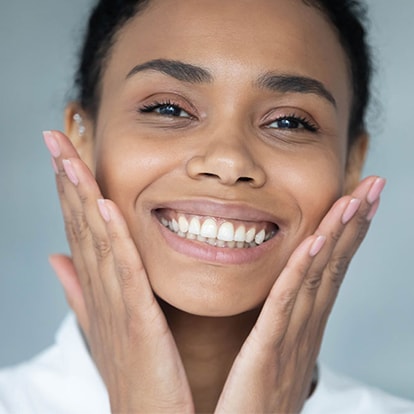 Nahaufnahme einer Frau, die in die Kamera lächelt und beide Hände an die Wange hält