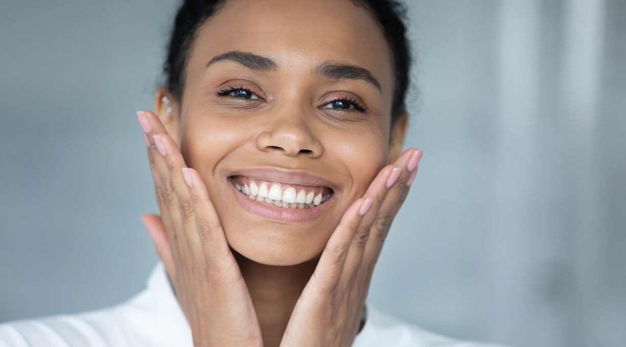 Nahaufnahme einer Frau, die in die Kamera lächelt und beide Hände an die Wange hält
