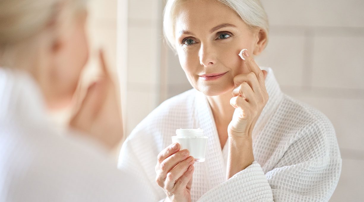 Reife Frau im Bademantel pflegt ihr Gesicht mit Creme vor dem Spiegel