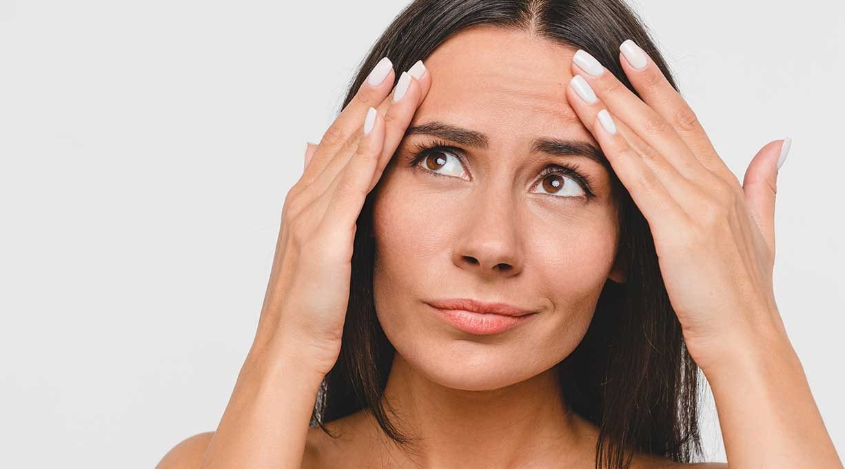 Junge Frau betrachtet besorgt ihre Stirn und fühlt feine Falten mit den Fingern.