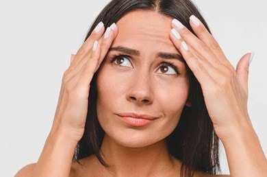 Junge Frau betrachtet besorgt ihre Stirn und fühlt feine Falten mit den Fingern.