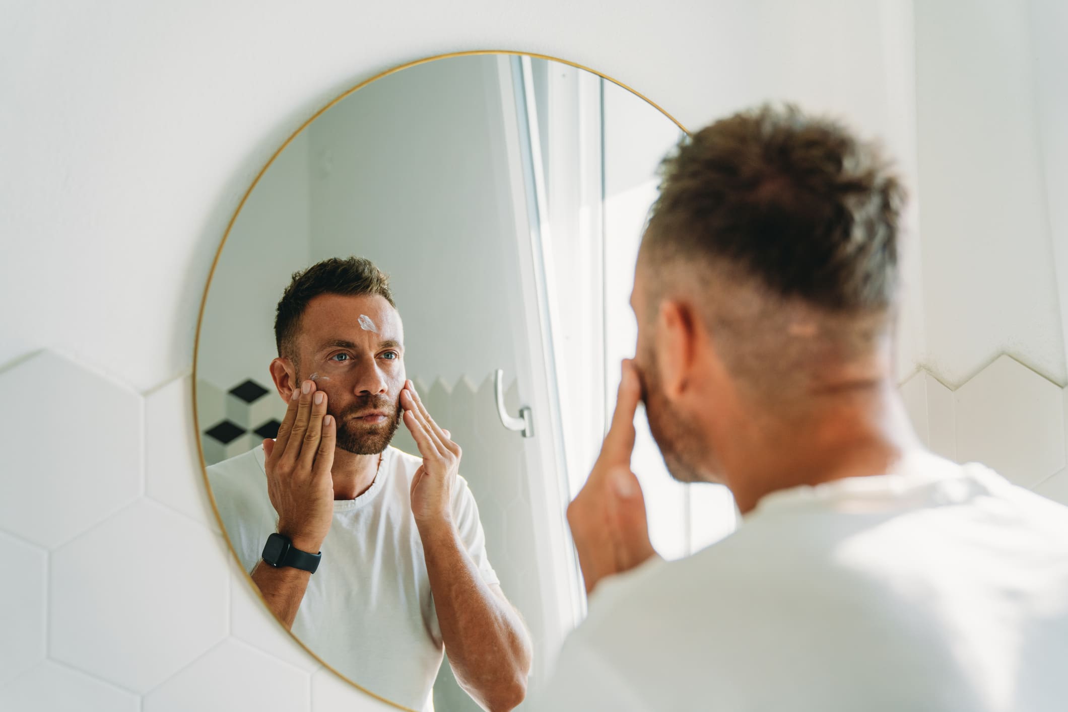 Ein Mann mit kurzen Haaren und leichtem Bart steht vor dem Spiegel und cremt sein Gesicht ein