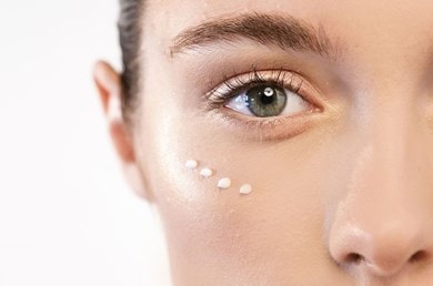 Eine Frau mit strahlend grünen Augen trägt sanft Augencreme unter ihren Augen auf.