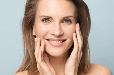 Eine Frau mit blonden Haaren vor blauem Hintergrund berührt sanft ihr Gesicht und lächelt