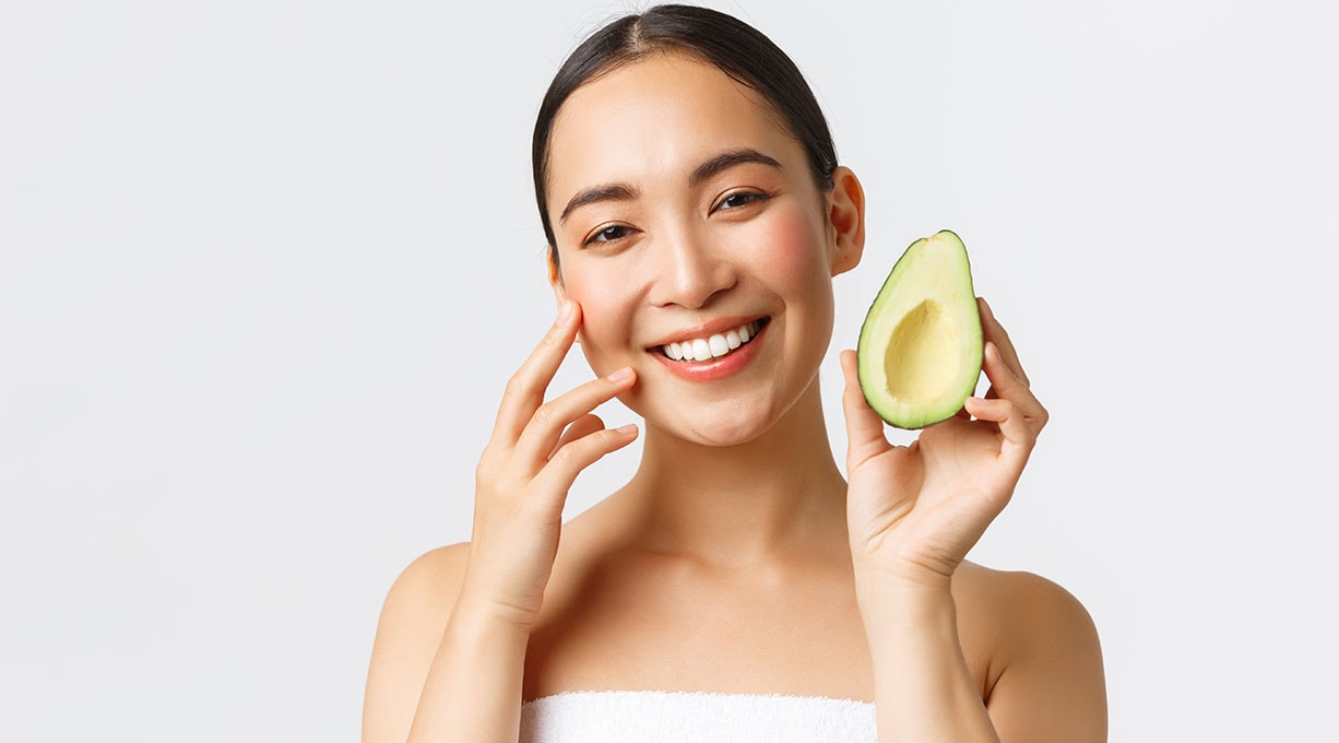 Junge Frau l&#228;chelt und ber&#252;hrt ihr Gesicht w&#228;hrend sie eine Avocado in der Hand h&#228;lt