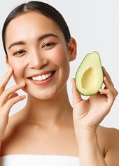 Junge Frau lächelt und berührt ihr Gesicht während sie eine Avocado in der Hand hält