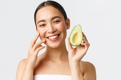 Junge Frau lächelt und berührt ihr Gesicht während sie eine Avocado in der Hand hält