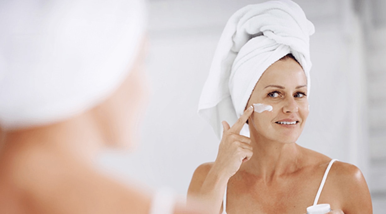 Frau mit Haarturban blickt in den Spiegel und cremt ihr Gesicht ein