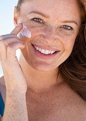 Frau mit roten langen Haaren steht vor dem Meer und cremt ihr Gesicht mit Sonnencreme ein