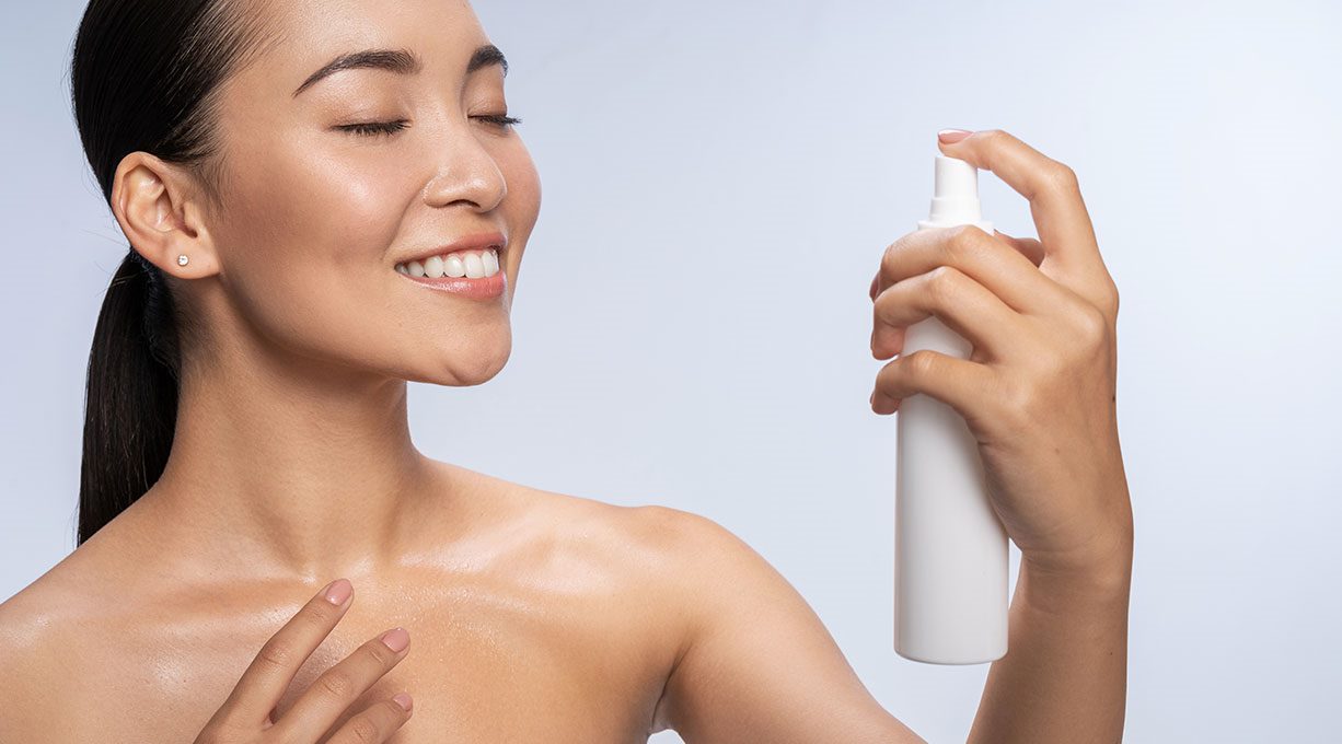 Eine junge, lächelnde Frau sprüht sich eine erfrischende Pflege auf Gesicht und Dekolleté. 