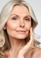 Eine Frau mittleren Alters mit graublonden Haaren und feinen Linien im Gesicht lächelt und streicht sanft über ihre linke Wange
