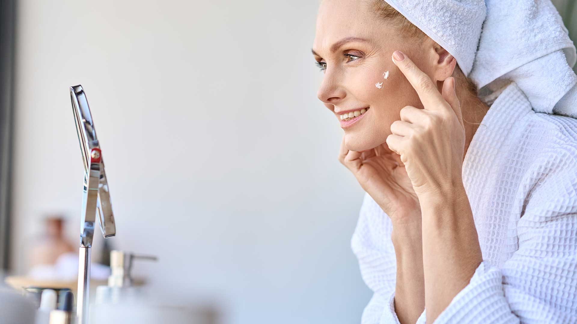 Eine Frau mittleren Alters mit Handtuchturban auf dem Kopf tupft sich mit dem Zeigefinger Gesichtspflege mit Kollagen-Peptiden auf die Haut