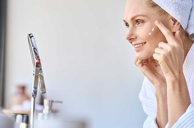 Eine Frau mittleren Alters mit Handtuchturban auf dem Kopf tupft sich mit dem Zeigefinger Gesichtspflege mit Kollagen-Peptiden auf die Haut