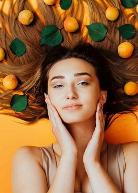Frau liegt auf orangenem Untergrund, ihre langen Haare sind von Aprikosen bedeckt