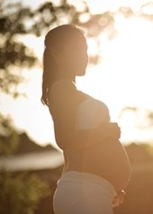 Eine schwangere Frau steht draussen während sie von der Sonne im Hintergrund angestrahlt wird