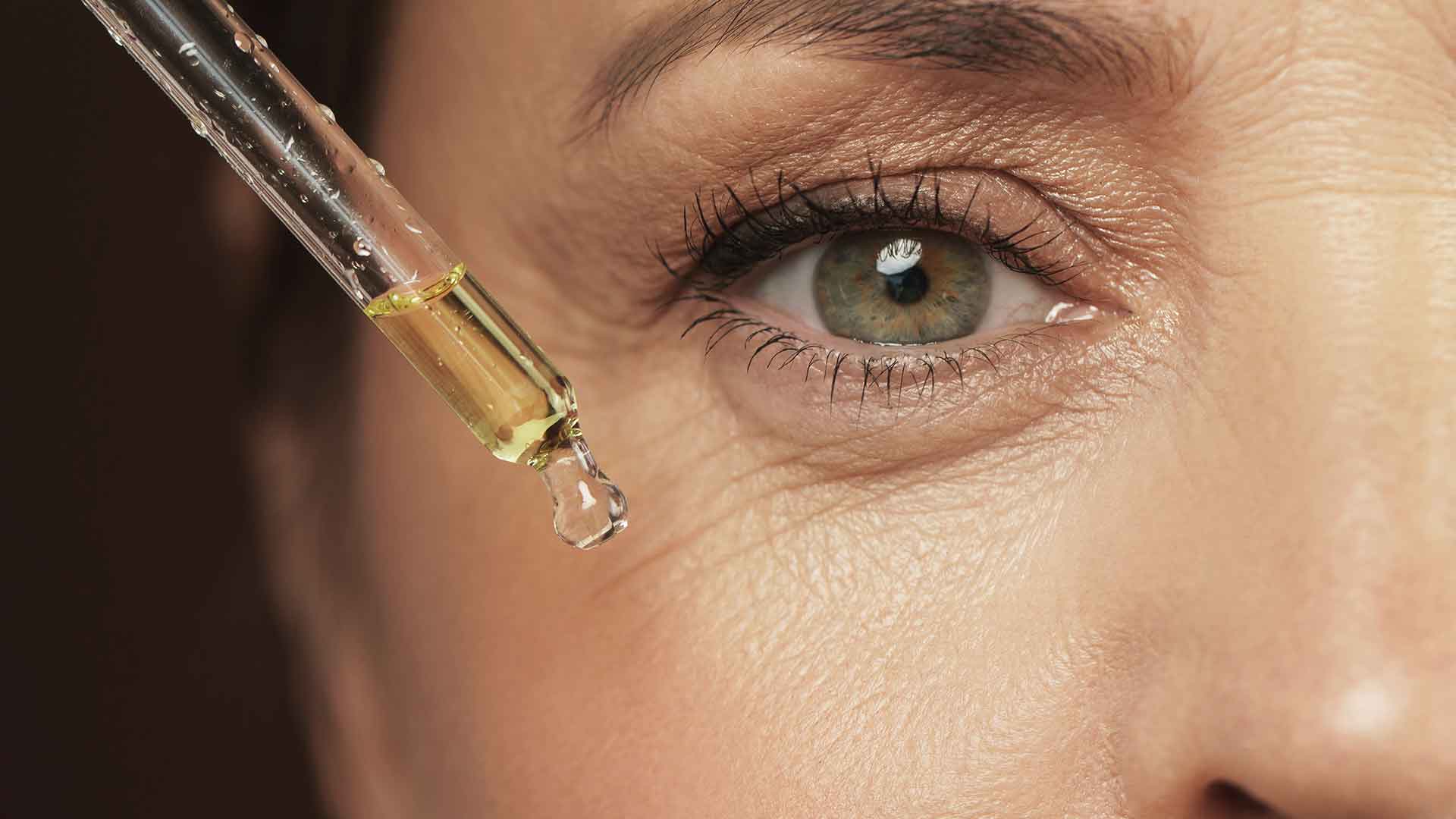 Nahaufnahme der Augenpartie einer Frau mit leichten Fältchen, die ein Serum aus einer Pipette auf die Gesichtspartie aufträgt