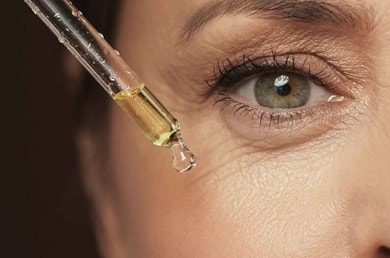 Nahaufnahme der Augenpartie einer Frau mit leichten Fältchen, die ein Serum aus einer Pipette auf die Gesichtspartie aufträgt