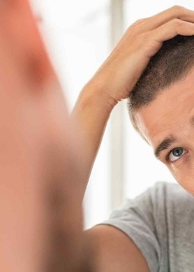 Eine Person betrachtet ihr Haar im Spiegel und fährt mit der Hand durch das kurze Haar. Sie trägt ein graues Oberteil.