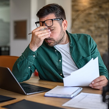Gestresster Mann am Schreibtisch mit Laptop und Dokumenten, hält sich die Nase