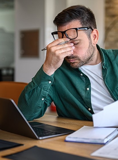 Gestresster Mann am Schreibtisch mit Laptop und Dokumenten, hält sich die Nase