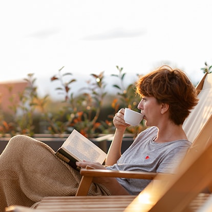 Frau entspannt auf der Terrasse, liest ein Buch und trinkt Kaffee bei Sonnenuntergang