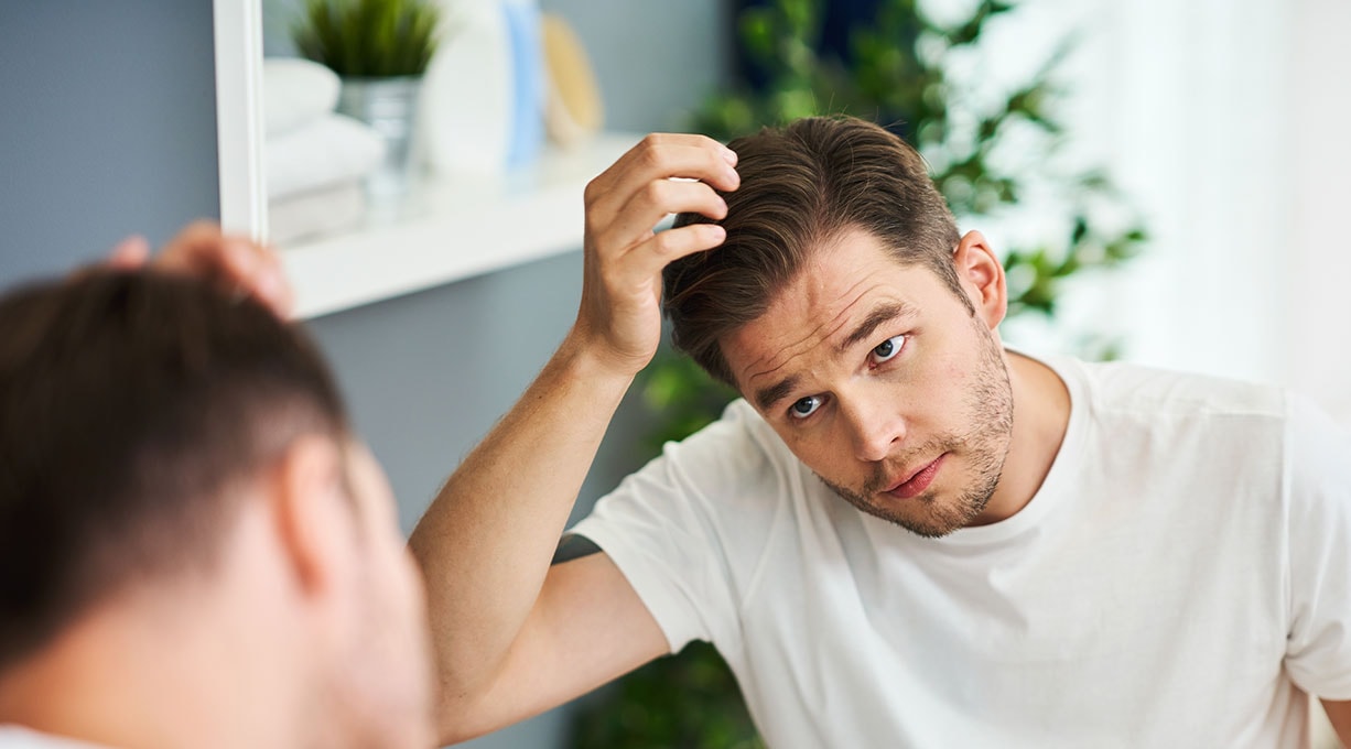 Ein Mann in weißem T-Shirt betrachtet sein Haar vor einem Spiegel in einem hellen Raum mit Pflanzen und Regalen im Hintergrund.