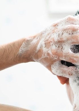 Une personne se lave les cheveux sous la douche, se masse les mains avec de la mousse dans les cheveux mouillés, gros plan de la tête vue de derrière.