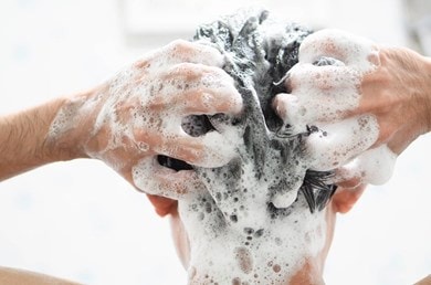 Une personne se lave les cheveux sous la douche, se masse les mains avec de la mousse dans les cheveux mouillés, gros plan de la tête vue de derrière.