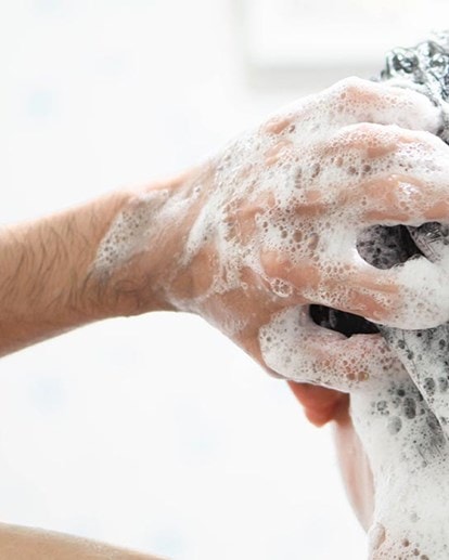 Une personne se lave les cheveux sous la douche, se masse les mains avec de la mousse dans les cheveux mouillés, gros plan de la tête vue de derrière.
