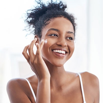 Junge lächelnde Frau trägt Creme auf ihre rechte Wange auf 