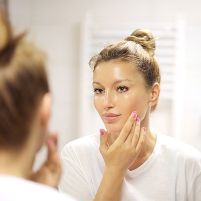 Junge Frau steht im Bad vor dem Spiegel und berührt ihre linke Wange