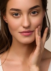 Jeune femme à la peau impeccable effleure doucement sa joue et regarde l'objectif, sur un fond neutre.