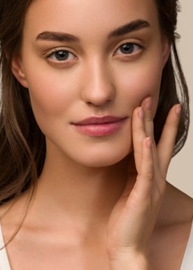 Jeune femme à la peau impeccable effleure doucement sa joue et regarde l'objectif, sur un fond neutre.