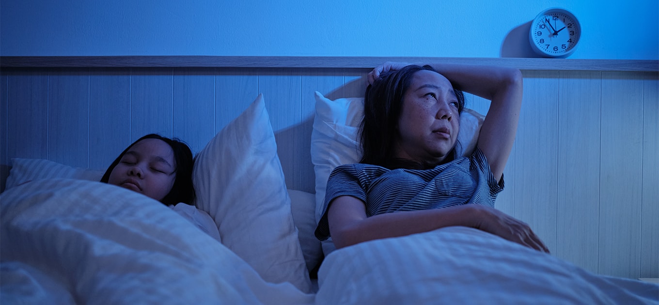Frau liegt nachts wach im Bett wegen Nachtschweiss neben einem schlafenden Kind