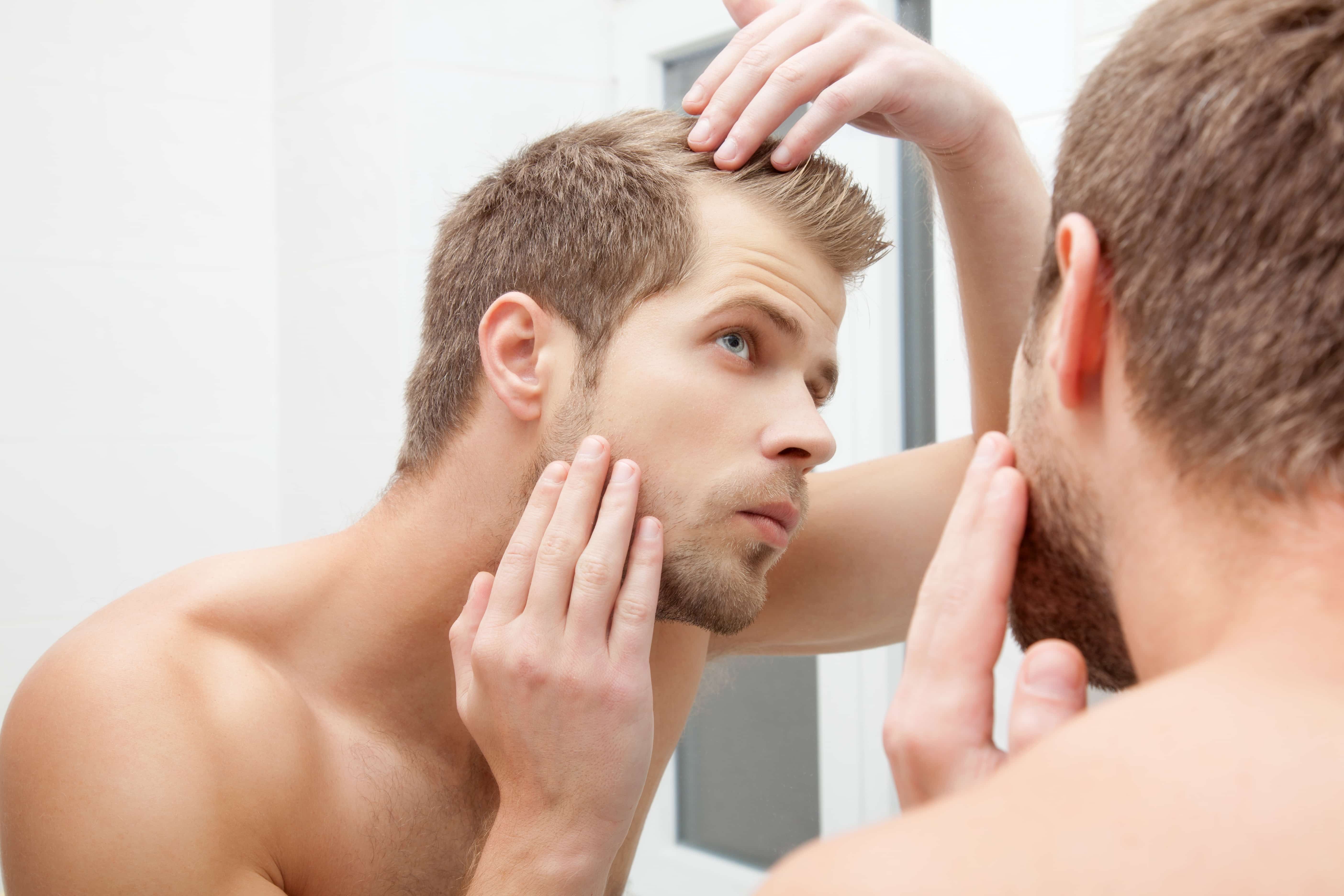 Junger Mann betrachtet im Spiegel kritisch seine Haare und Kopfhaut auf Anzeichen von Haarausfall.