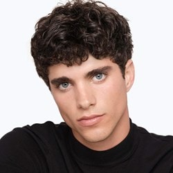 Haut du corps et visage d'un jeune homme portant un haut noir et ayant des cheveux  bruns bouclés.