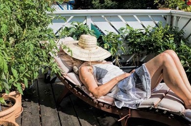 Une femme en robe d'été et chapeau de paille est allongée sur une chaise longue sur une terrasse en bois, entourée de plantes, et profite du soleil.
