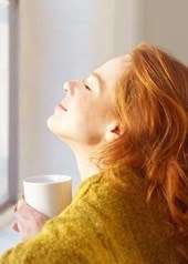 Eine Frau sitzt mit geschlossenen Augen und einer Tasse vor einem geschlossenen Fenster und lässt die Sonne in ihr Gesicht scheinen